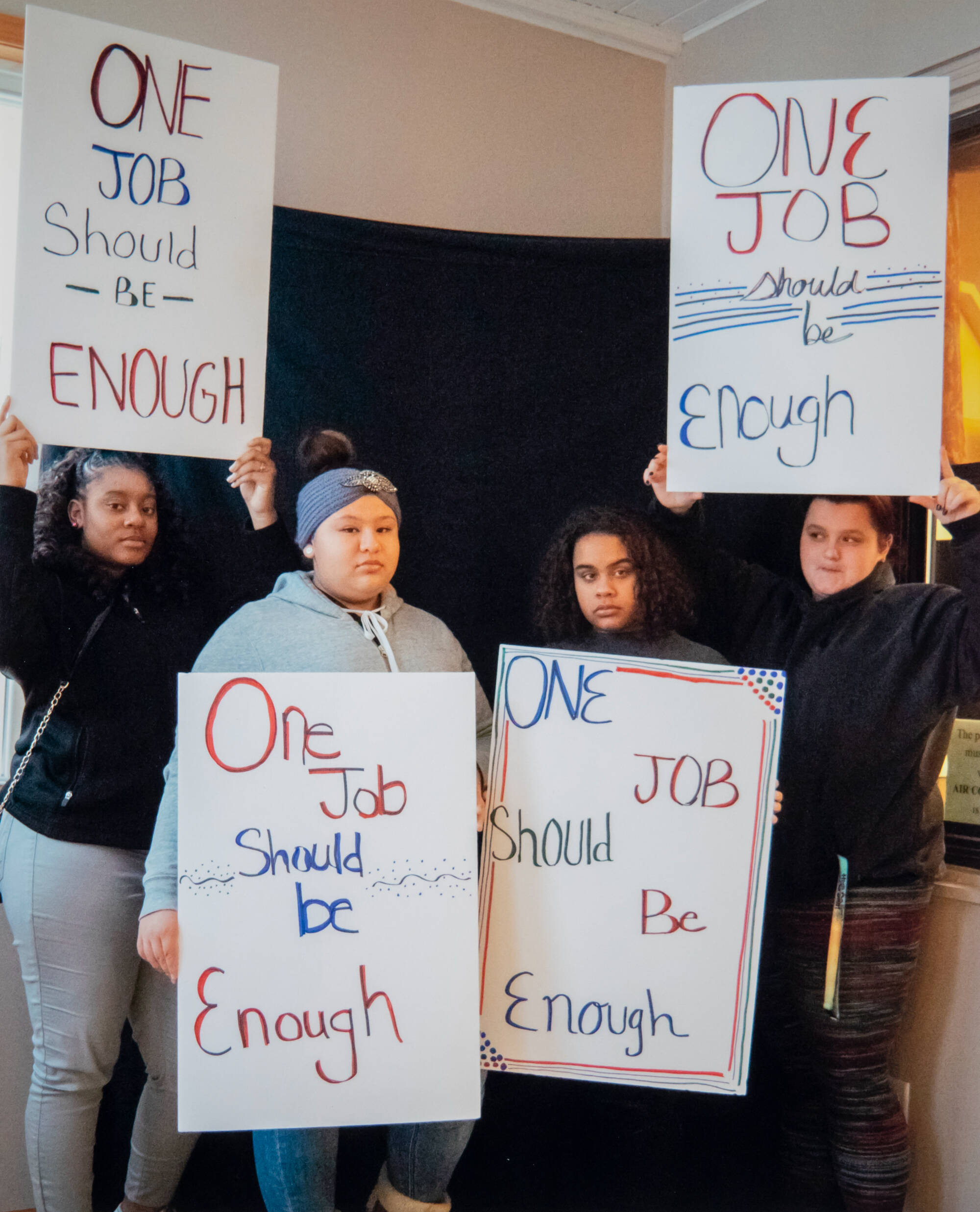 Color photograph of four people standing together, holding signs that say "One Job Should Be Enough."